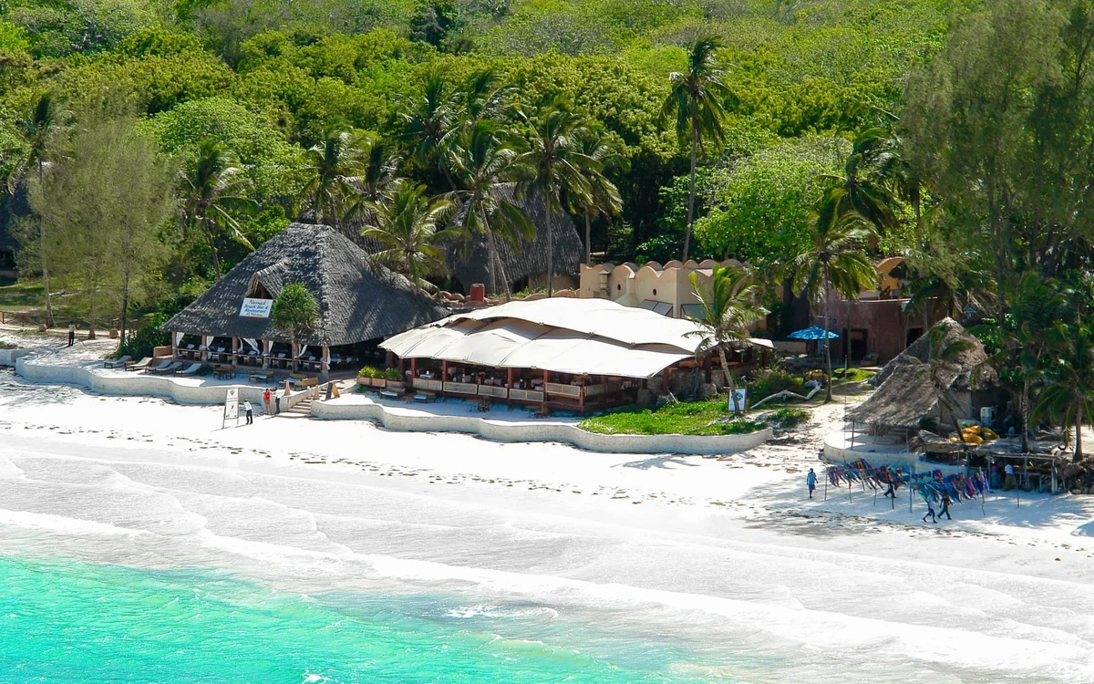 Beachfront view of The Sands at Nomad resort with white sand, turquoise water, palm trees, and open-air seaside restaurant in Diani Beach, Kenya.